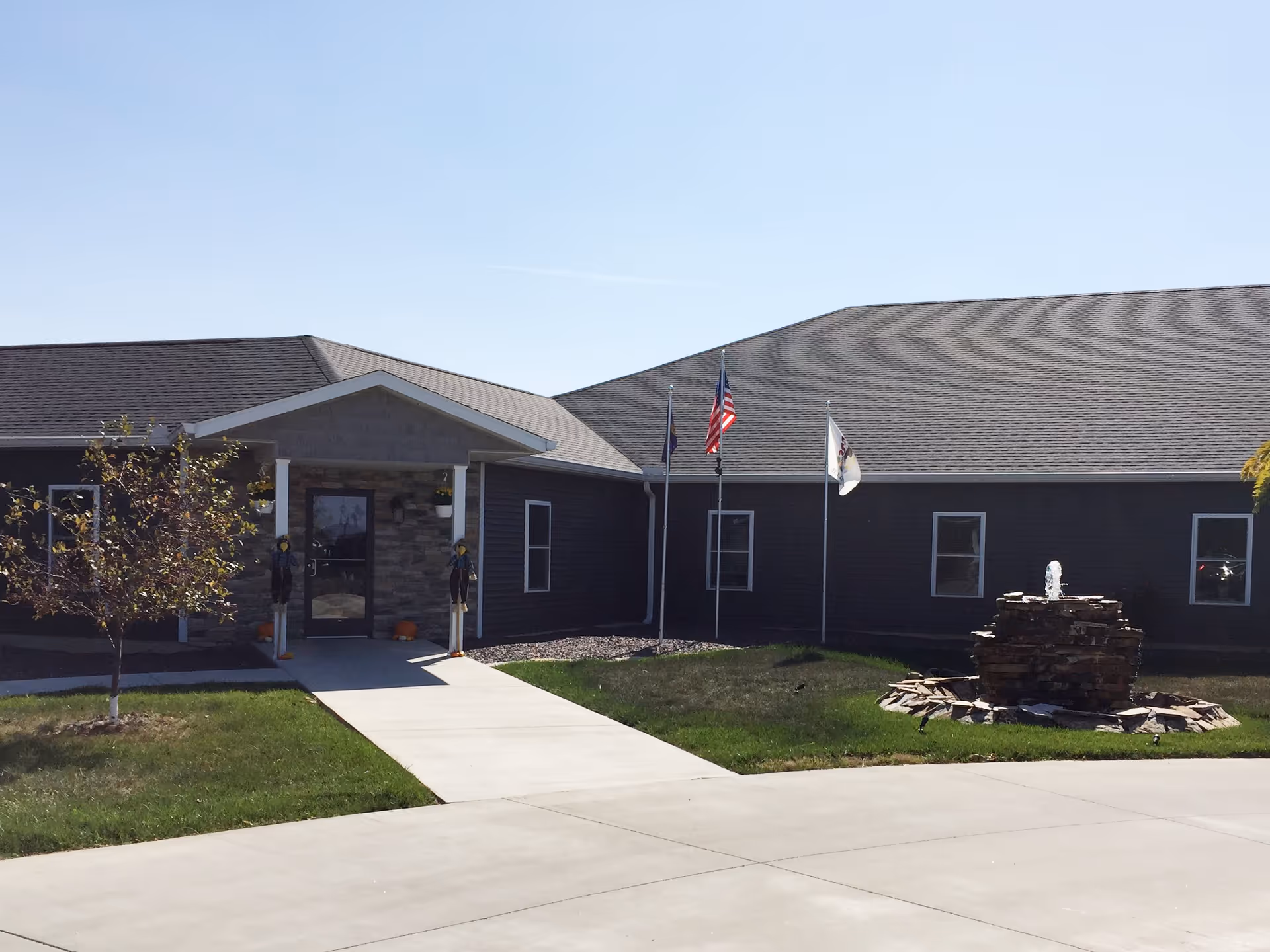 Exterior view of a single-story building with a stone entrance, three flagpoles with flags, a small tree, and a stone water fountain on a grassy lawn under a clear blue sky.