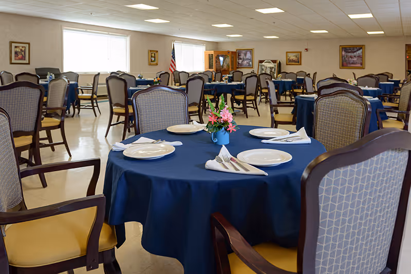 A spacious dining room with multiple round tables covered in dark blue tablecloths. Each table is set with white plates, napkins, and silverware. There are cushioned chairs with wooden frames around the tables. The room has large windows letting in natural light, framed pictures on the walls, and an American flag in the background.