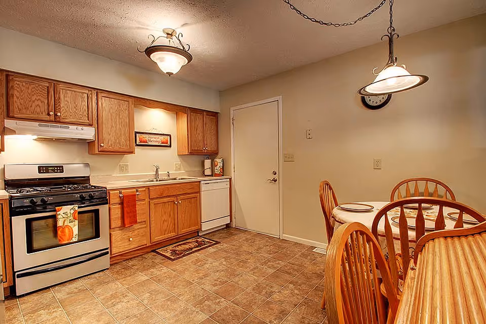 A kitchen and dining area featuring wooden cabinets, a stainless steel gas stove with an oven, a white dishwasher, and a sink. The kitchen has a tiled floor and a small dining table with four wooden chairs set with plates. There are two ceiling lights, one above the kitchen area and one hanging above the dining table. A door is visible in the background.