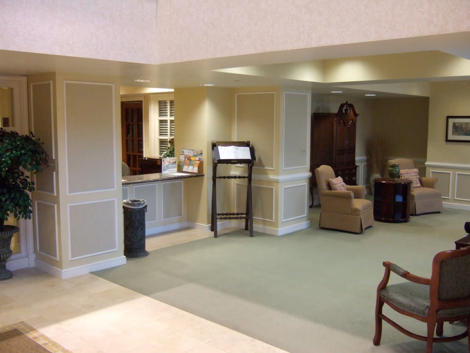 Interior view of a senior living facility lobby area with beige walls and green carpet. There is a reception desk with brochures, a wooden stand holding an open book, two beige armchairs with pillows around a small round wooden table, and a wooden cabinet in the background. A potted plant is visible on the left side.