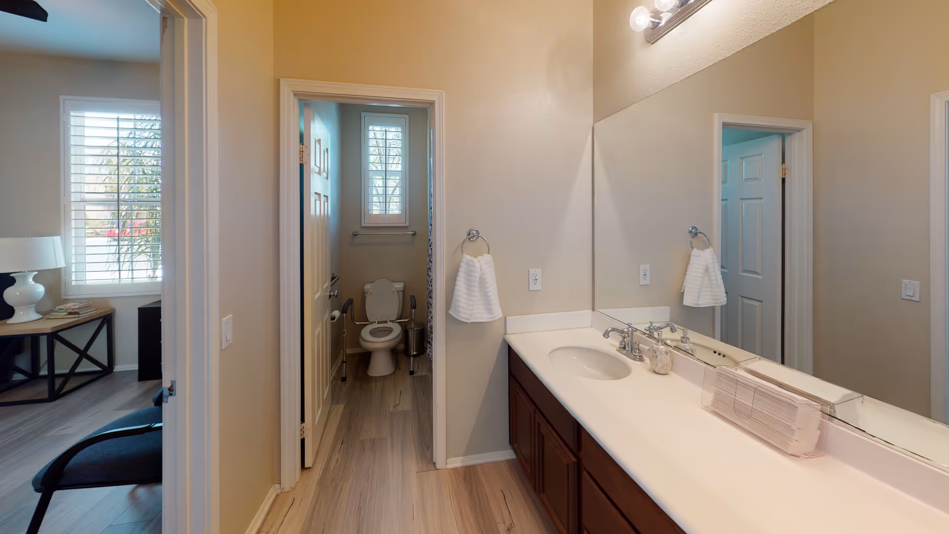 Interior view of a bathroom area in a senior living facility. The image shows a long countertop with a sink, a large mirror above it, and a hand towel hanging on a ring. Through an open door, a toilet with safety rails is visible, along with a small window with blinds. Adjacent to the bathroom is a room with a window, a table with books, and a lamp.