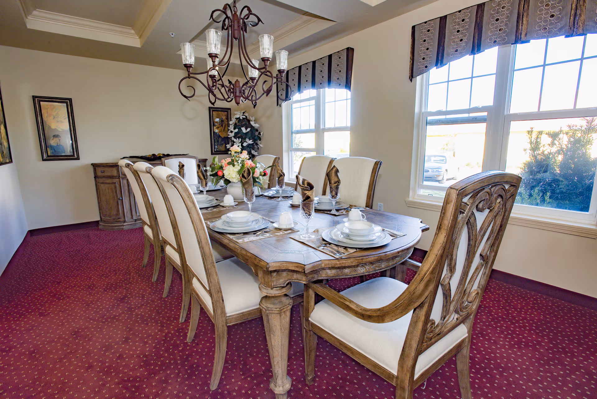 A formal dining room with a wooden dining table set for six people. The table has white plates, cups, glasses, and brown folded napkins. There is a floral centerpiece on the table. The room has beige walls, red carpet with a dotted pattern, two framed pictures on the wall, a wooden sideboard, and large windows with valances letting in natural light.
