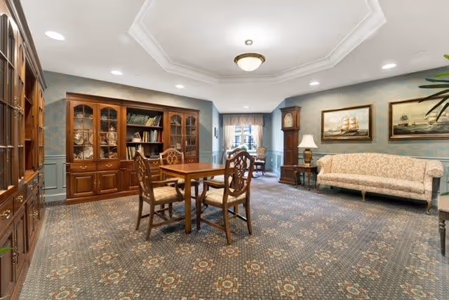 A spacious, traditionally decorated common area with a wooden table and four chairs in the center. There is a large wooden cabinet with glass doors filled with books and decorative items along the left wall. A grandfather clock and a side table with a lamp are positioned near a beige upholstered sofa on the right. Two framed paintings of ships hang on the wall above the sofa. The room has patterned carpet flooring and a ceiling light fixture.