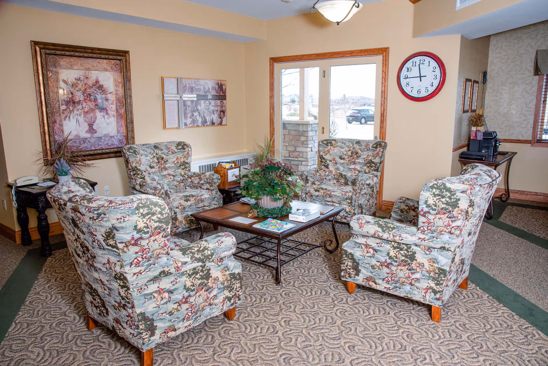 A cozy sitting area with four patterned armchairs arranged around a rectangular coffee table with a plant centerpiece. The room has beige walls with framed artwork, a large window with a view outside, a wall clock, and a small table with a printer in the corner.