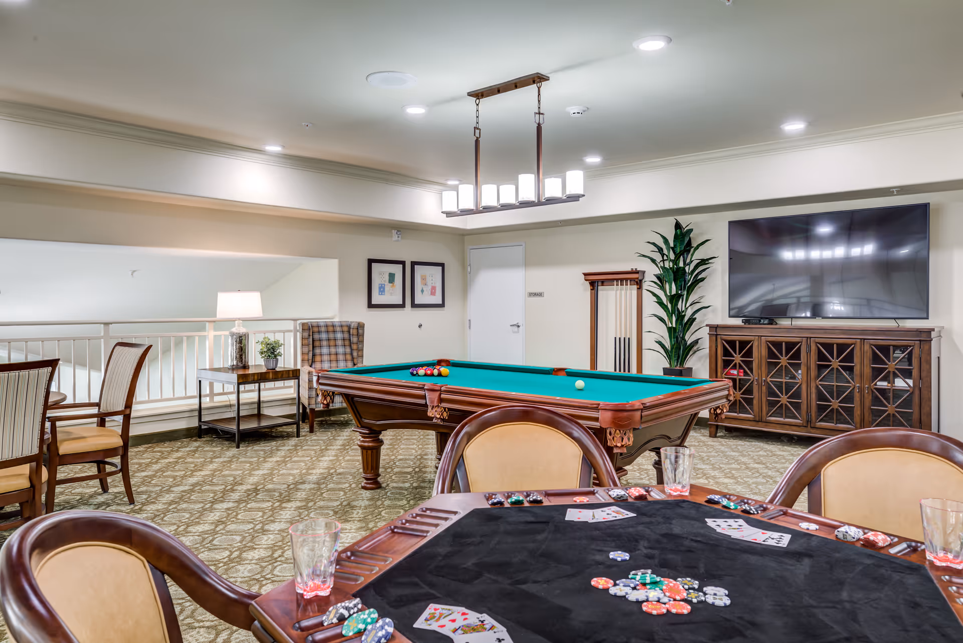 A recreational room featuring a pool table with balls arranged for a game, a poker table with playing cards and poker chips, several chairs, a large flat-screen TV mounted on a wooden cabinet, a potted plant, and framed artwork on the wall. The room has carpeted flooring and is well-lit with ceiling lights and a hanging light fixture above the pool table.