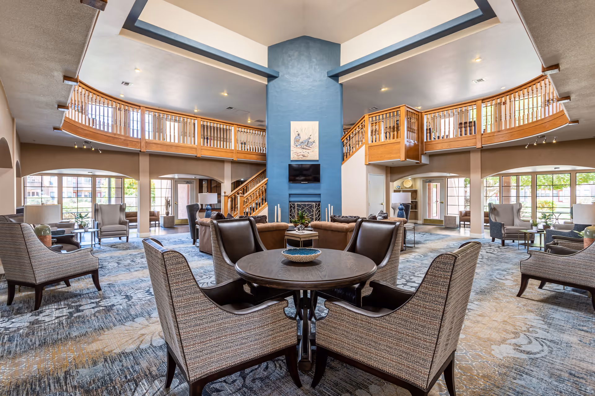 Spacious senior living facility common area with a high ceiling and a mezzanine balcony. The room features a central blue fireplace with a TV mounted above it, surrounded by comfortable seating including armchairs and sofas. Large windows allow natural light to fill the space, and a round table with four chairs is in the foreground.
