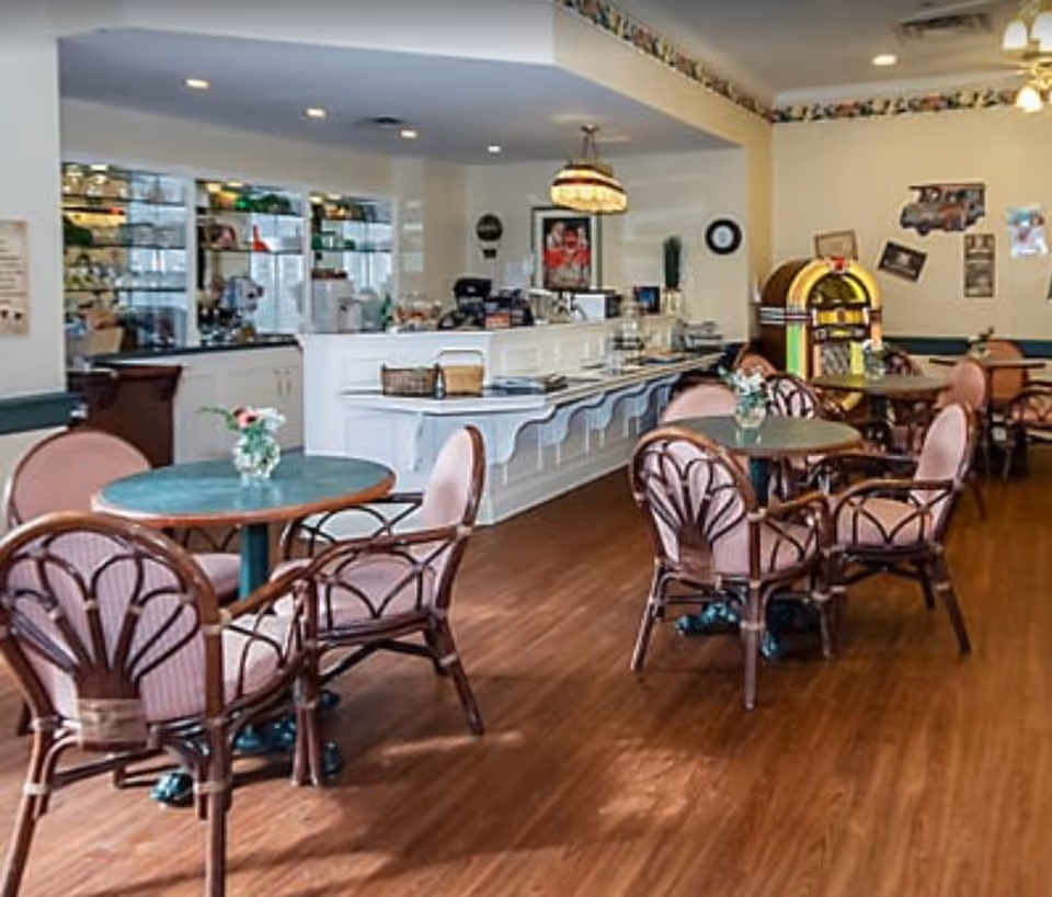 Bright dining room with round tables and pink upholstered chairs, a white counter/bar and a jukebox in the background.