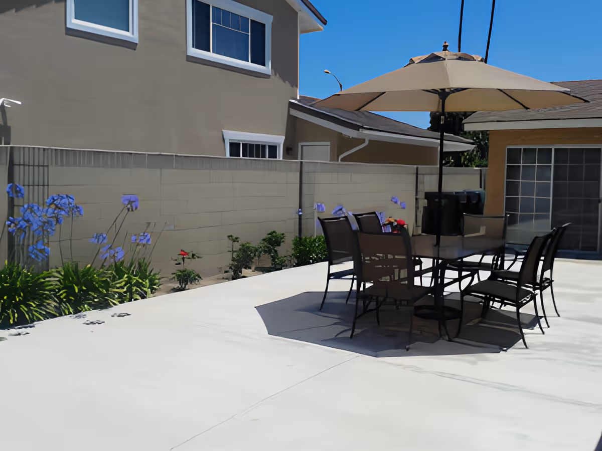 Outdoor patio area with a table and six chairs under a large beige umbrella. The patio is surrounded by a beige wall with some plants and flowers along the edge. The sky is clear and blue.
