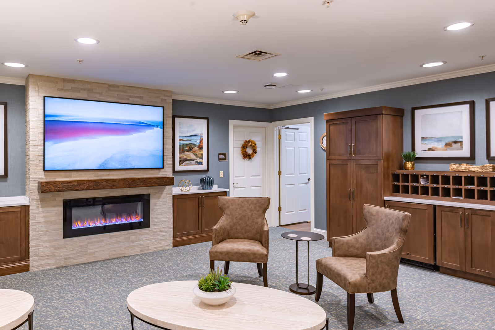 A cozy senior living facility common area with two brown upholstered chairs, a round coffee table with a small plant, a wall-mounted flat screen TV above a modern electric fireplace, wooden cabinets, and framed artwork on the walls.