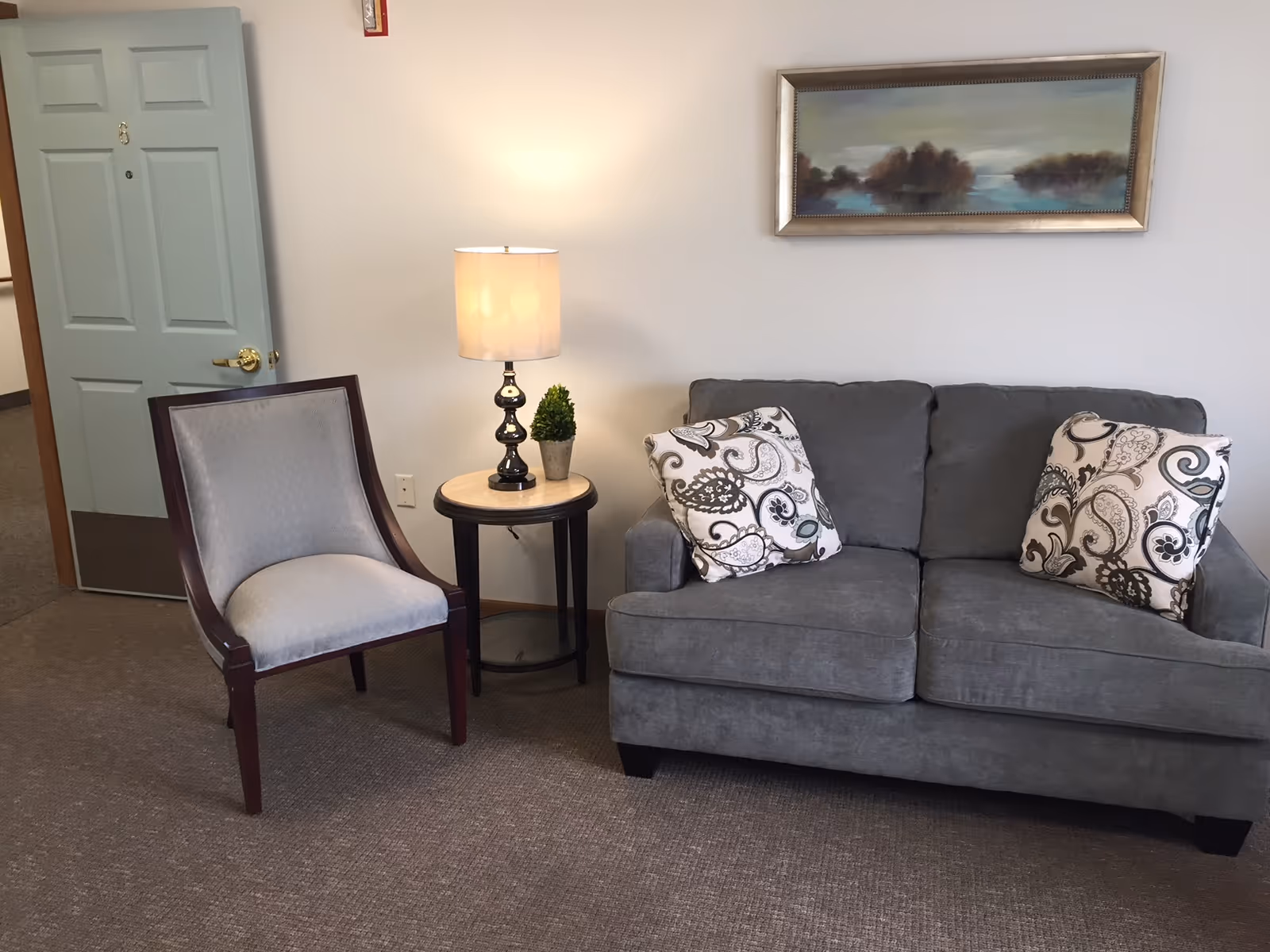 A cozy seating area with a gray upholstered sofa adorned with two patterned pillows, a wooden chair with light gray upholstery, a small round side table with a lamp and a small potted plant, and a framed landscape painting on the wall above the sofa.