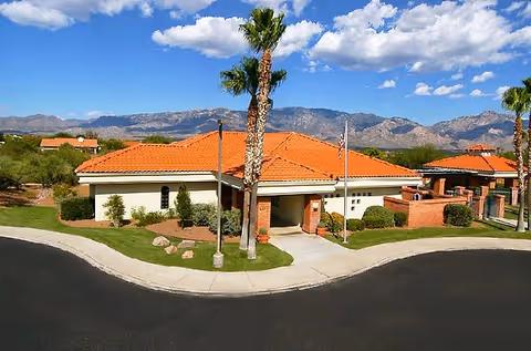 Single-story building with a red tile roof and palm trees in front, set against mountains under a blue sky.