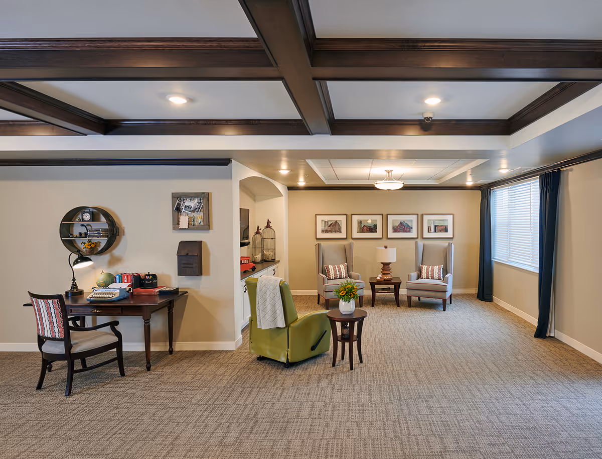 A spacious living room area with beige walls and carpeted floor. The ceiling has dark wooden beams and recessed lighting. On the left, there is a wooden desk with a chair, a desk lamp, a globe, and books. Above the desk is a round wall shelf and a small bulletin board. In the center, there is a green recliner chair with a white blanket draped over it and a small round wooden side table with a vase of flowers. In the background, two gray armchairs with patterned cushions flank a wooden side table with a lamp. Four framed pictures hang on the wall behind the armchairs. A large window with dark curtains is on the right side of the room.