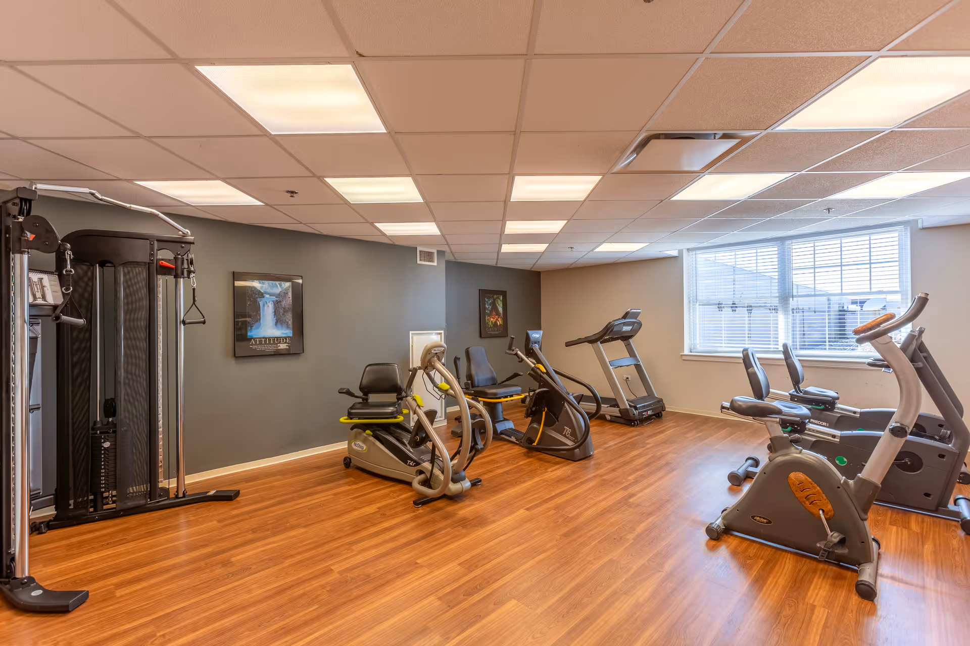 A fitness room with exercise equipment including stationary bikes, a treadmill, and a multi-function weight machine. The room has wood flooring, a drop ceiling with fluorescent lights, and a large window letting in natural light. Motivational posters are hung on the walls.