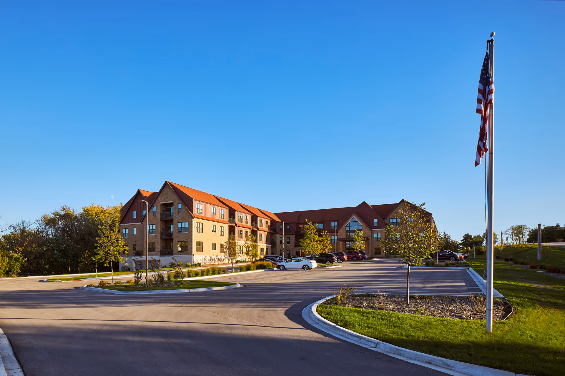Exterior view of a multi-story senior living facility building with a red roof and beige walls, surrounded by a parking lot with several cars and a flagpole with an American flag in the foreground under a clear blue sky.