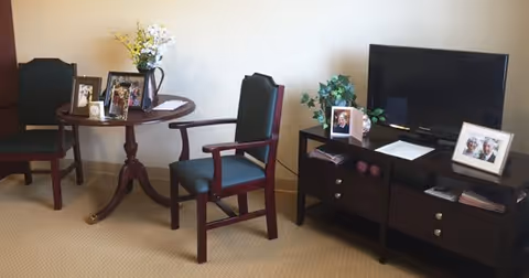 A cozy interior room with two wooden chairs with green cushions, a small round wooden table holding a vase with flowers and several framed photos, and a dark wooden TV stand with a flat-screen television, more framed photos, and decorative items.