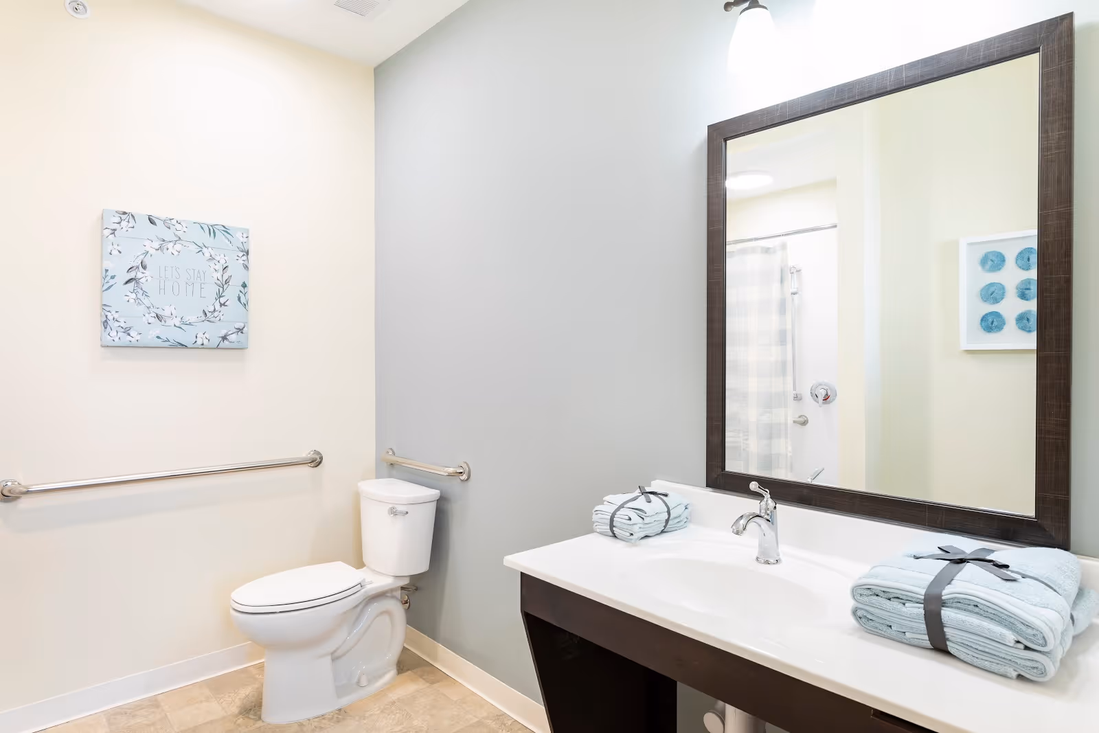 Bright accessible bathroom with a toilet, grab bars, a sink and mirror, and neatly folded towels on the counter.