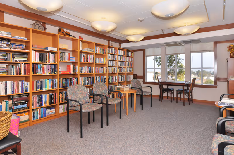 Communal library/reading room with wall-to-wall bookshelves, several chairs and tables, and large windows.