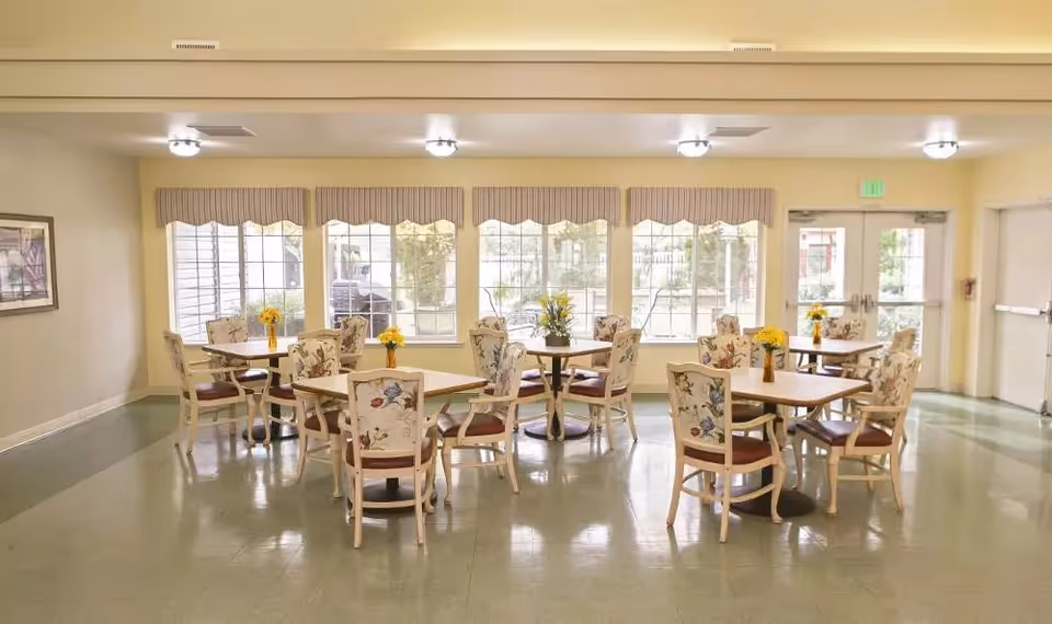 Bright communal dining room with several round tables and floral-upholstered chairs arranged near large windows and doors.