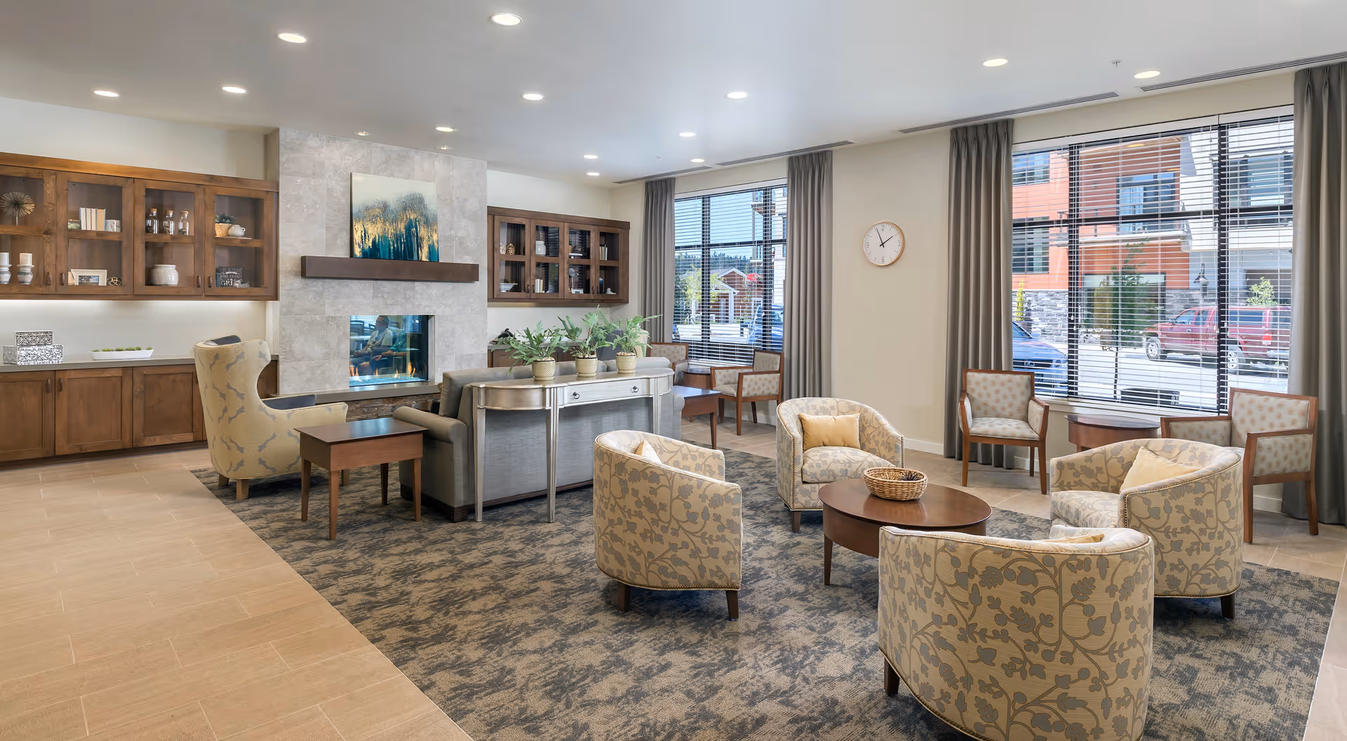 A bright and spacious senior living community lounge area with patterned armchairs arranged around a round wooden coffee table on a carpeted floor. There is a gray sofa with a console table behind it, potted plants, a modern fireplace with artwork above it, wooden cabinets with glass doors, large windows with blinds and curtains, and a wall clock.