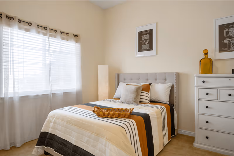 A cozy bedroom with a bed featuring a striped bedspread in shades of white, black, and orange, adorned with several pillows. Next to the bed is a tall white dresser with a large amber glass bottle on top. The room has beige walls, a window with sheer curtains allowing natural light to enter, and a tall floor lamp in the corner. Two framed artworks hang above the bed.