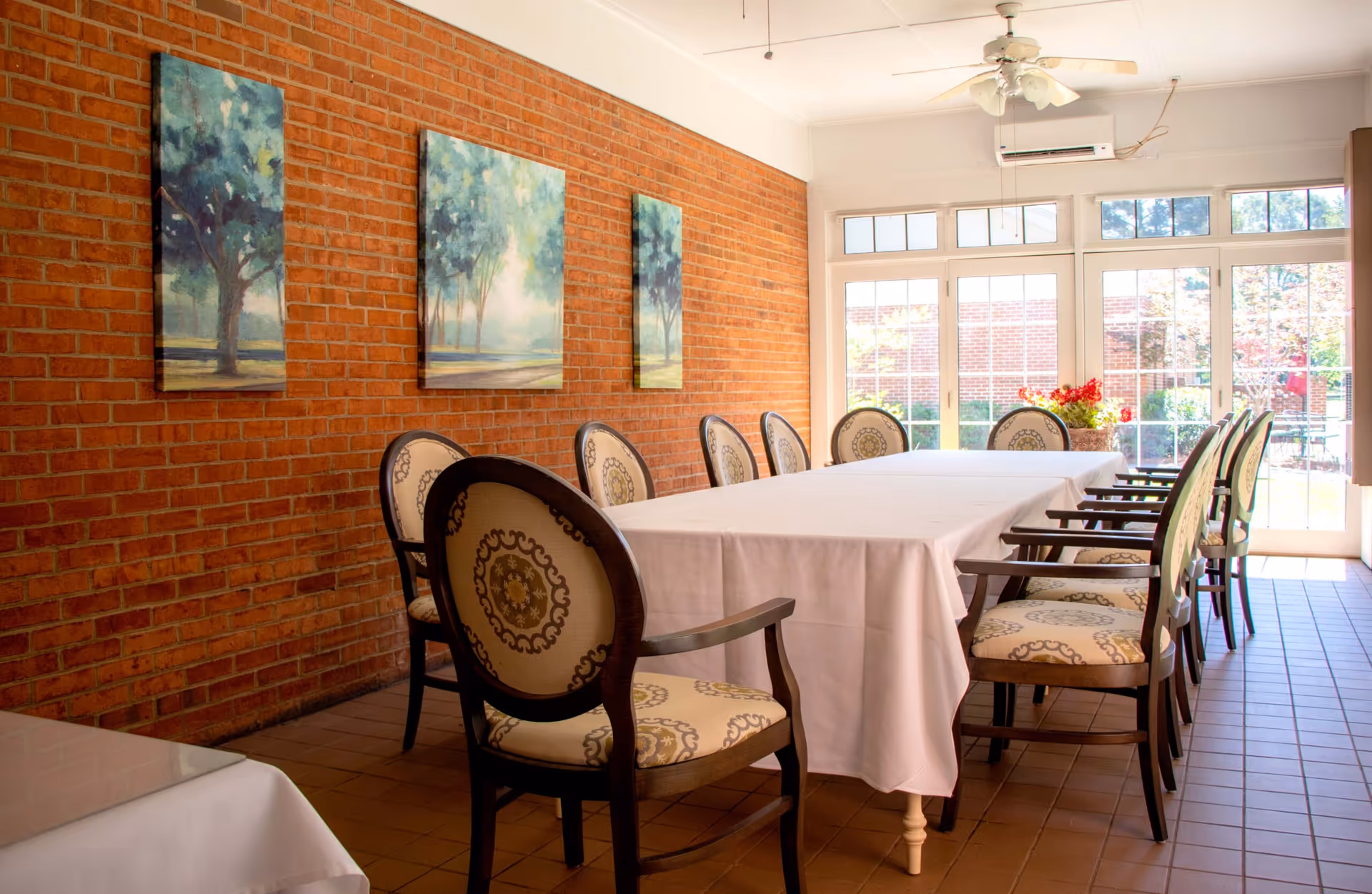 A bright dining room with a long rectangular table covered with a white tablecloth, surrounded by cushioned chairs with ornate backs. The room has a brick wall adorned with three paintings of trees, large windows and glass doors letting in natural light, and a ceiling fan above.
