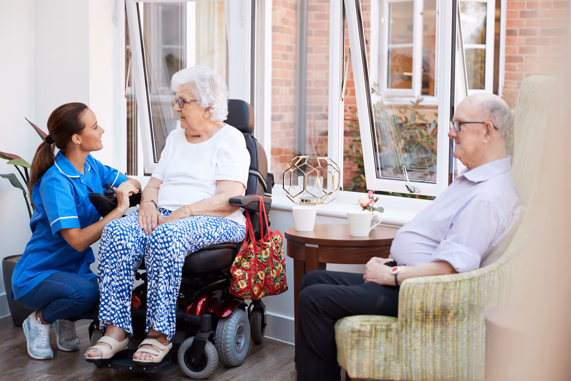 A female caregiver in a blue uniform kneels and talks to an elderly woman sitting in a motorized wheelchair near a window. An elderly man sits in a green armchair nearby, looking towards them. The room has large open windows, a small round wooden table with decorative items, and a potted plant in the corner.