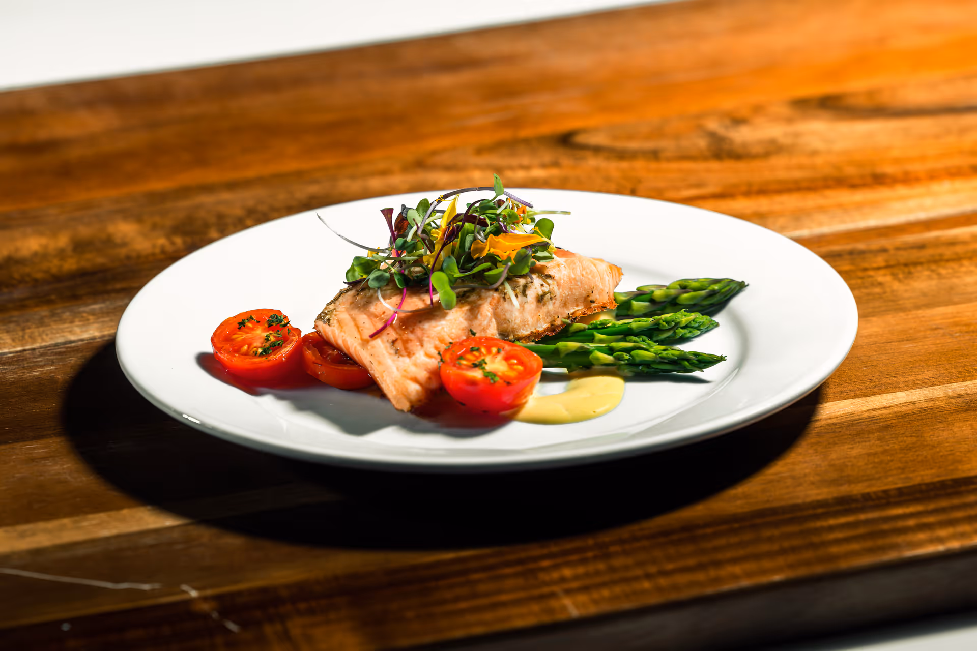 A white plate with a serving of cooked salmon topped with microgreens, accompanied by asparagus spears, halved cherry tomatoes, and a small dollop of yellow sauce, placed on a wooden table.