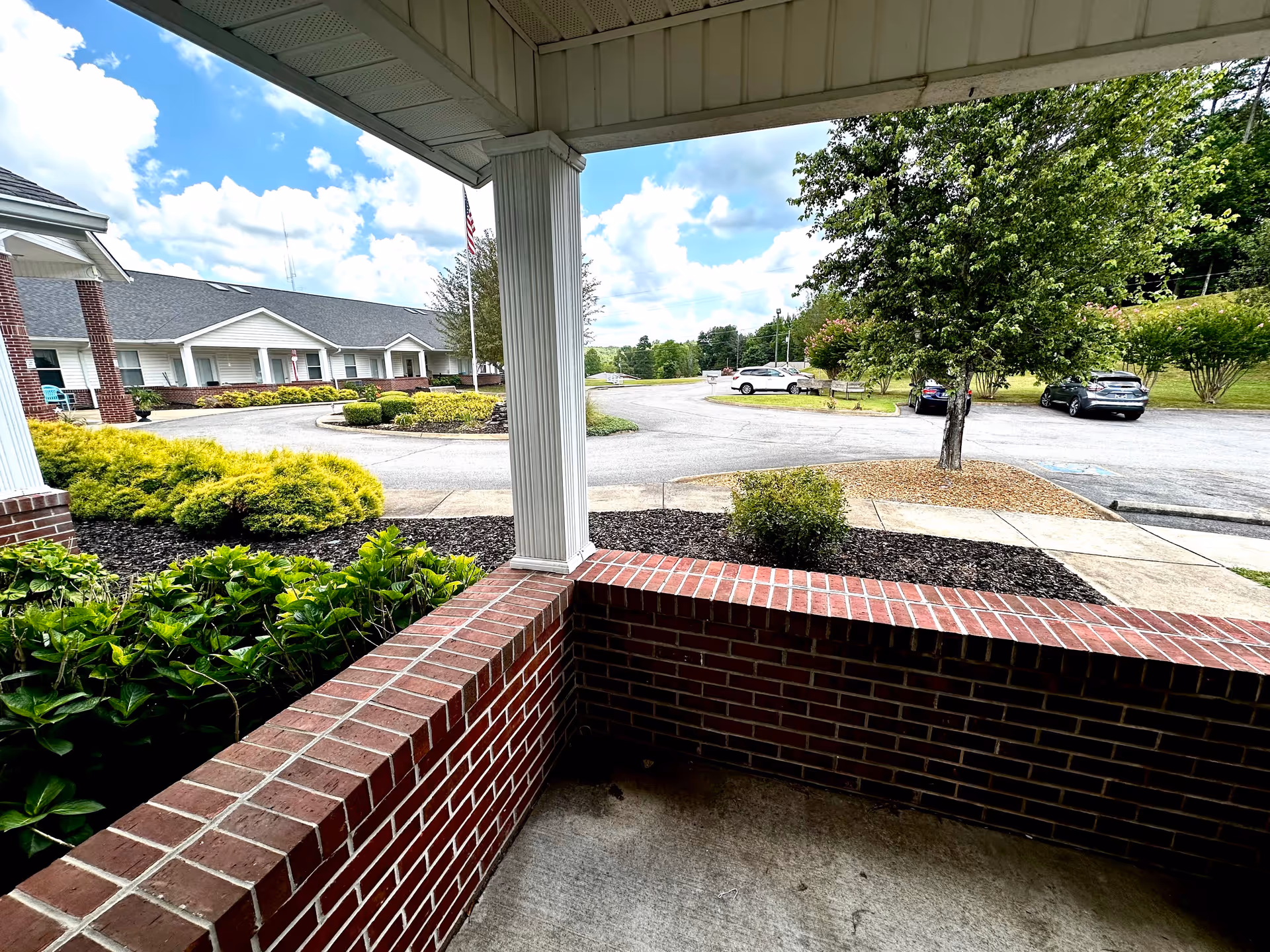 View from a covered porch area with red brick walls looking out onto a parking lot and landscaped garden area with bushes and a tree. In the background, there is a single-story building with white siding and red brick columns under a partly cloudy sky.