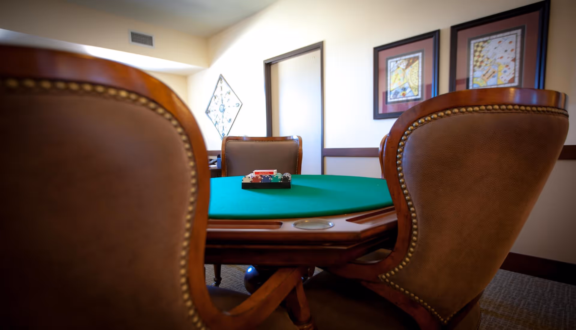 A card table with a green felt surface surrounded by brown leather chairs with wooden frames. On the table, there is a tray holding poker chips and a deck of cards. The room has beige walls with framed artwork and a door in the background.