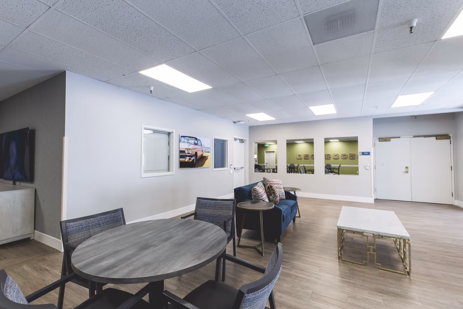 A modern common area in a senior living facility featuring a round table with four chairs, a blue sofa with decorative pillows, a small round side table, and a rectangular coffee table with a marble top and gold-colored legs. The walls are light gray with three rectangular windows looking into another room with green walls and additional seating. A flat-screen TV is mounted on the left wall above a white cabinet. The ceiling has recessed lighting panels, and the floor is covered with wood-style flooring.