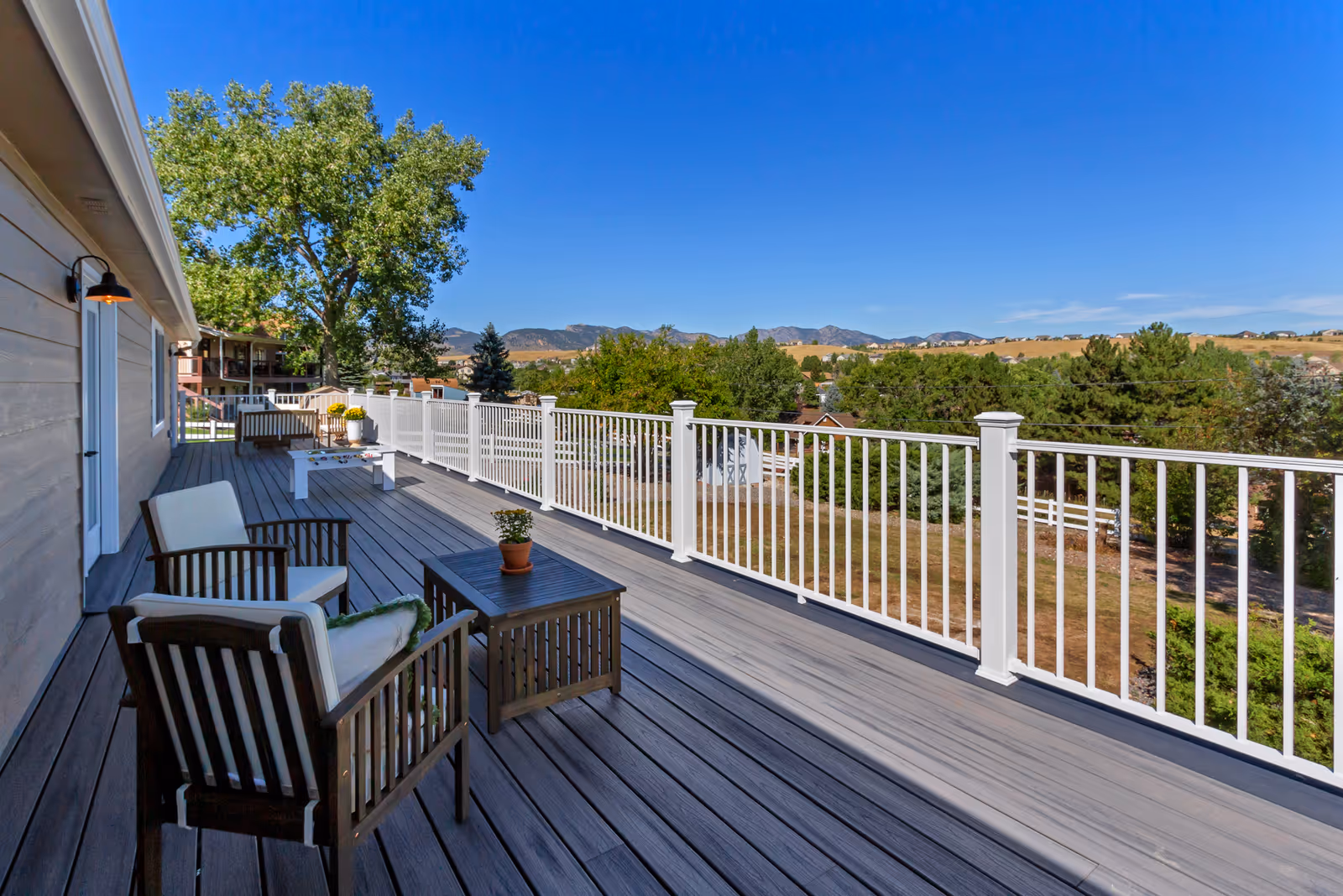 A spacious outdoor balcony with wooden flooring and white railing, furnished with cushioned chairs and small tables with potted plants. The balcony overlooks a scenic view of trees, houses, and distant hills under a clear blue sky.