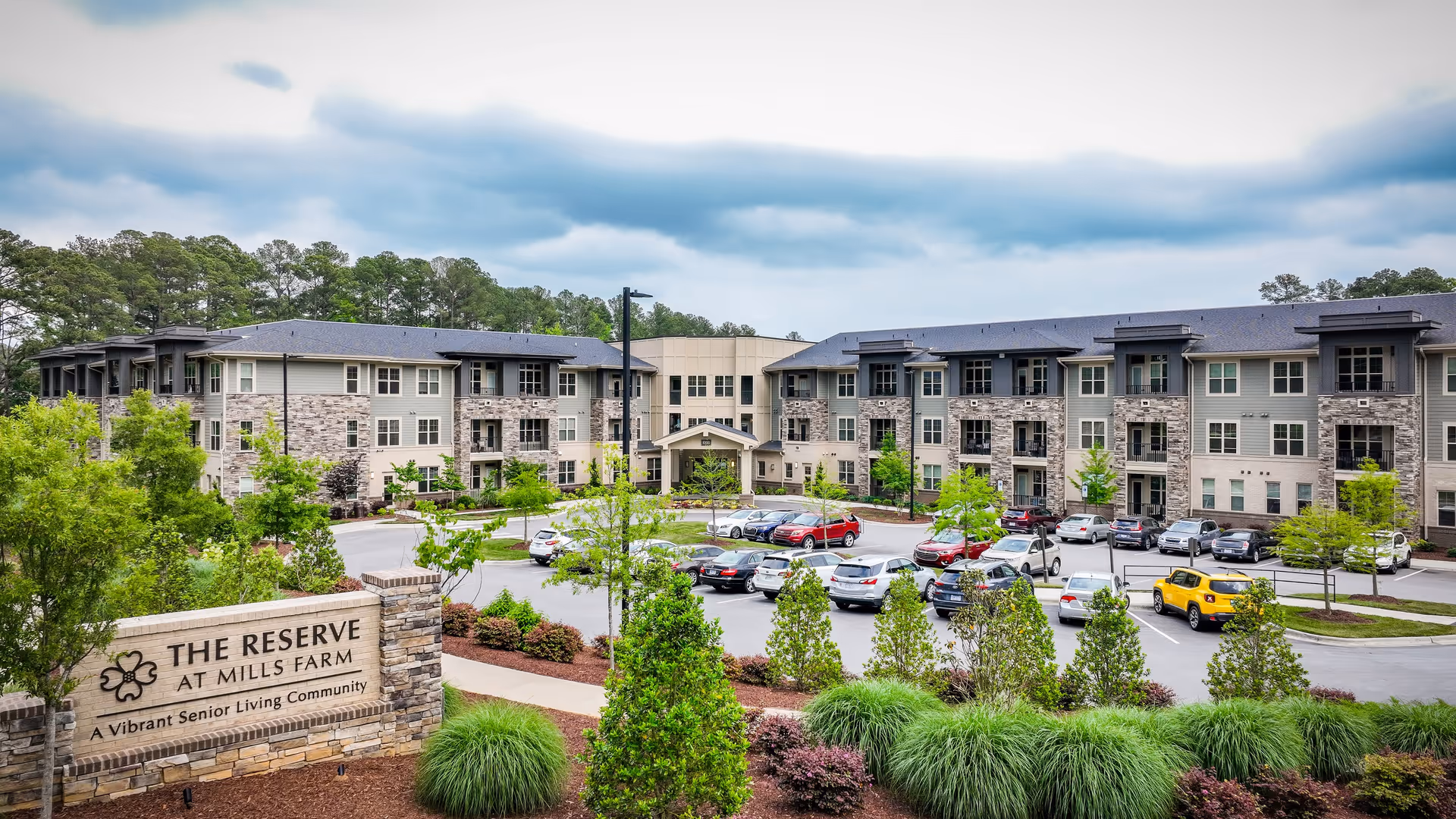 Exterior view of The Reserve at Mills Farm senior living community showing a large three-story building with balconies, a parking lot with several cars, landscaped greenery, and a sign in the foreground that reads 'The Reserve at Mills Farm, A Vibrant Senior Living Community'.