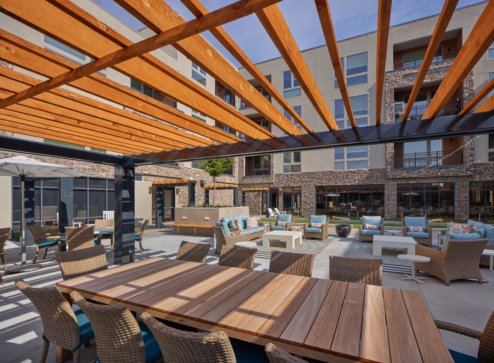 Outdoor seating area at The 501 at Mattison Estate featuring a wooden pergola overhead, a large wooden table with wicker chairs, and multiple cushioned lounge chairs and sofas arranged around coffee tables on a concrete patio, with a multi-story building in the background.