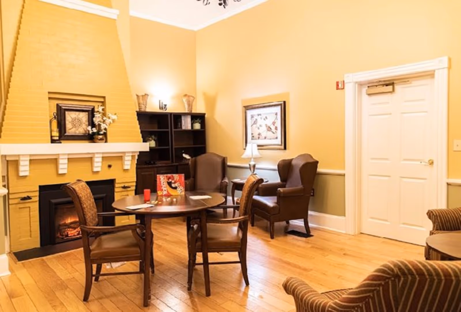 A cozy living room with a yellow brick fireplace, a round wooden table with four chairs, two brown leather armchairs, a bookshelf, a framed picture on the wall, and a white door. The room has warm yellow walls and wooden flooring.