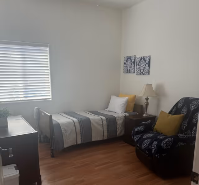 A small bedroom with a single bed covered in a striped gray and white blanket, a white pillow, and a yellow pillow. Next to the bed is a wooden nightstand with a lamp. There is a dark patterned armchair with a yellow pillow on it. Two decorative wall hangings are above the bed. A window with closed blinds is on the left wall, and a wooden dresser with a small plant on top is partially visible. The floor is wooden.