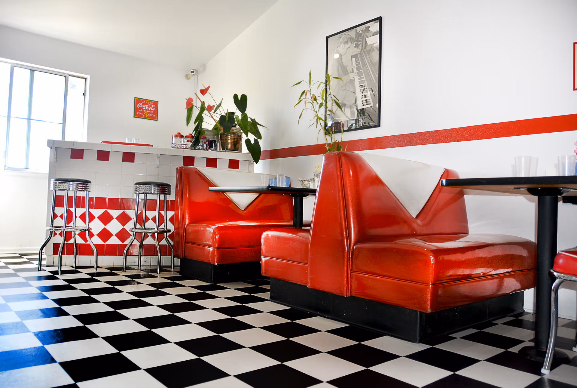 A retro-style dining area with red vinyl booth seating, black and white checkered floor tiles, and a small counter with chrome bar stools. The walls are white with a red stripe and decorated with plants and a framed black and white photo.