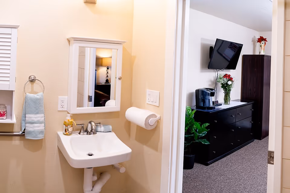 A small bathroom area with a white sink, a wall-mounted mirror cabinet, a towel ring holding a blue and white striped hand towel, and a soap dispenser. To the right, an open doorway leads to a room with a black dresser, a mounted flat-screen TV, a coffee maker, a vase with flowers, and a tall dark cabinet.