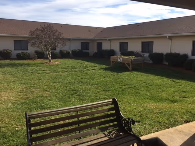 A sunny courtyard with a wooden bench in the foreground, a grassy lawn, a raised planter, and single-story building windows around the perimeter.