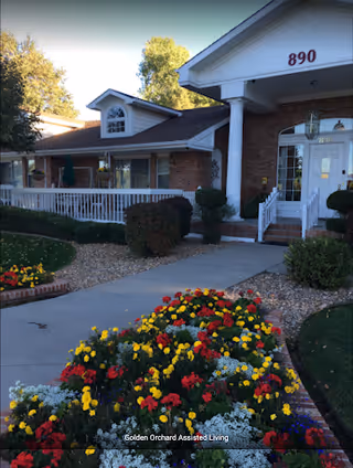 Exterior view of a brick building with white columns and a porch railing, surrounded by landscaped gardens with colorful flowers and bushes. The building has the number 890 displayed above the entrance.