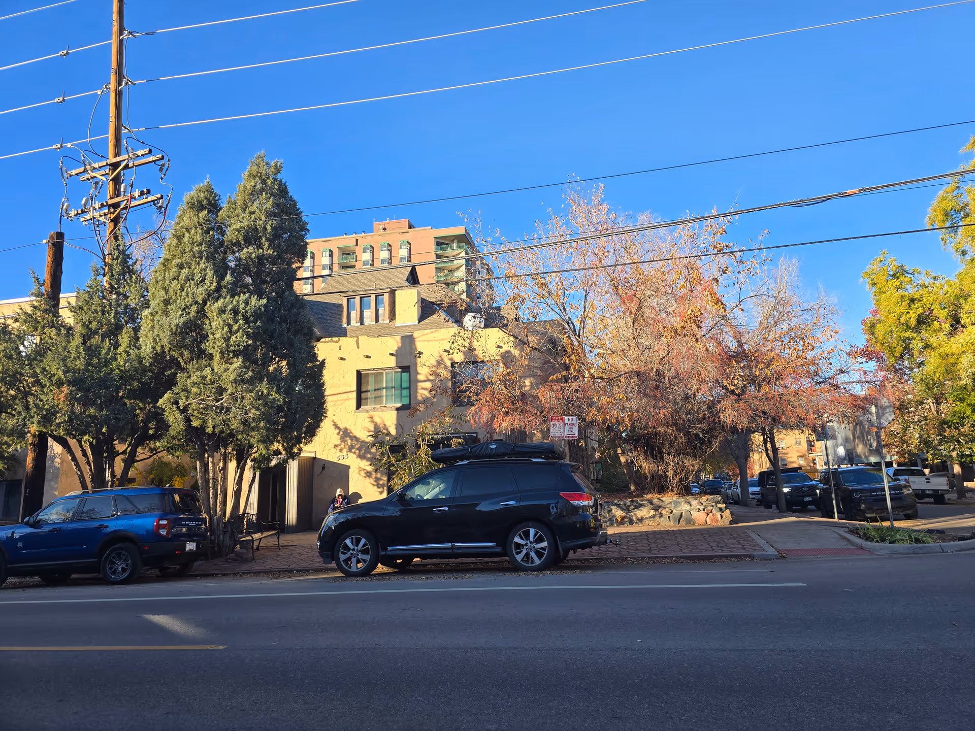Street view of a building with trees and parked cars in front under a clear blue sky. The building has a beige exterior with multiple windows and a small entrance. Several cars are parked along the street, and there are power lines overhead.