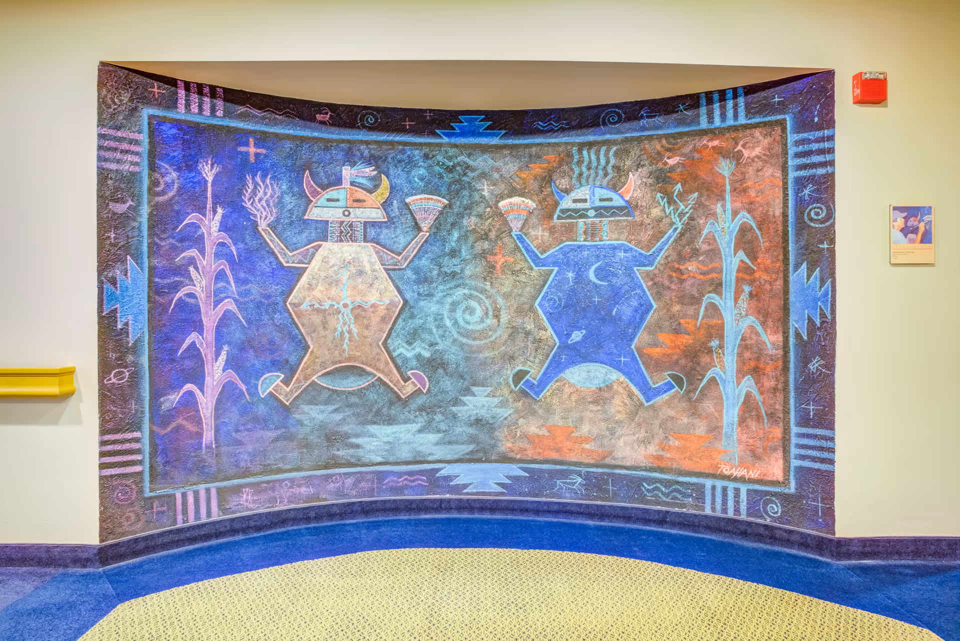 Colorful Native-style mural on a curved interior wall depicting two stylized horned figures and cornstalks in a hallway.
