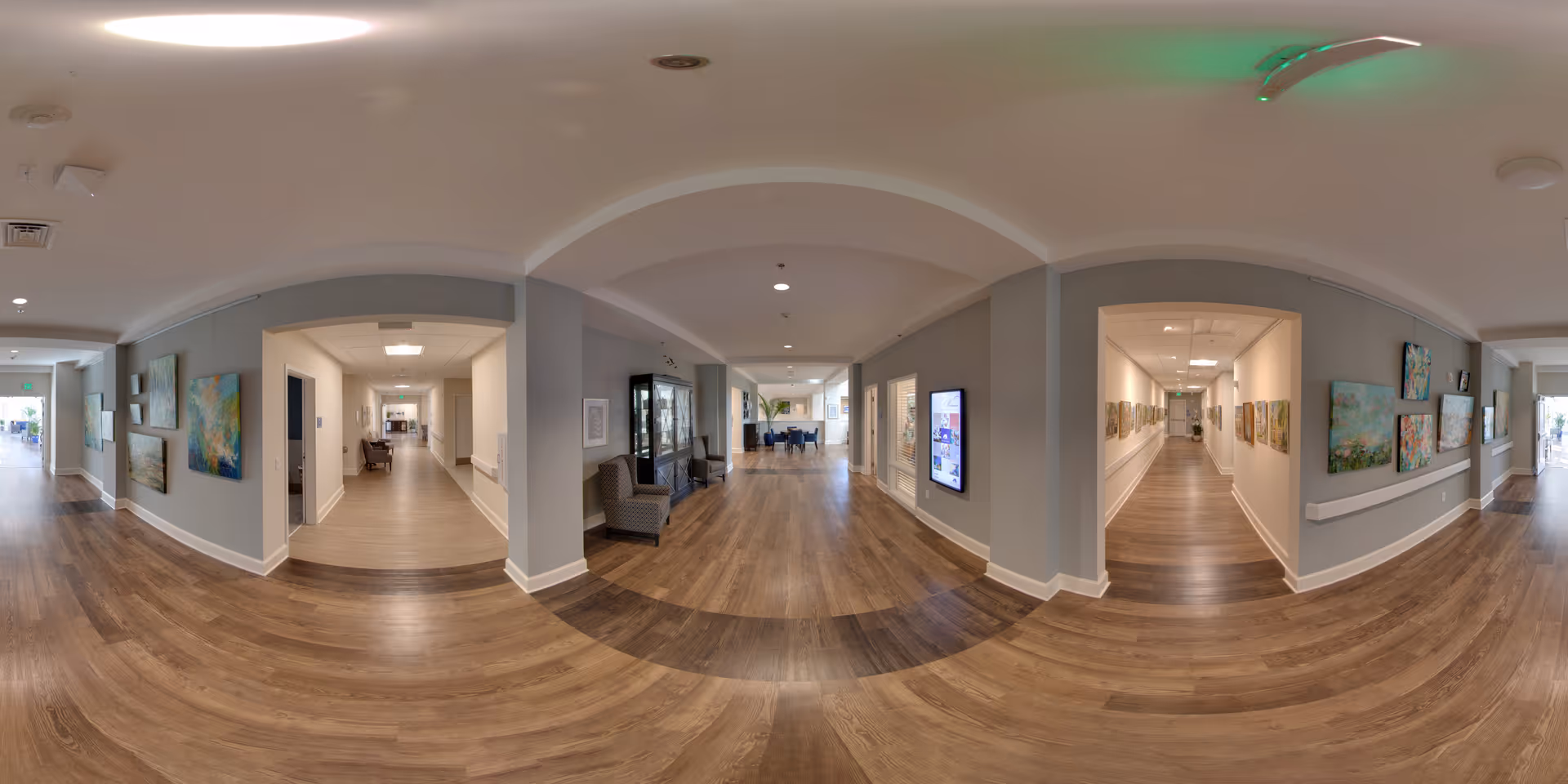 A wide hallway in Indigo Hall Assisted Living and Memory Care with wooden flooring and light gray walls adorned with colorful paintings. There are seating areas with chairs and a display cabinet along the hallway. The space is well-lit with ceiling lights and has multiple doorways leading to other rooms.
