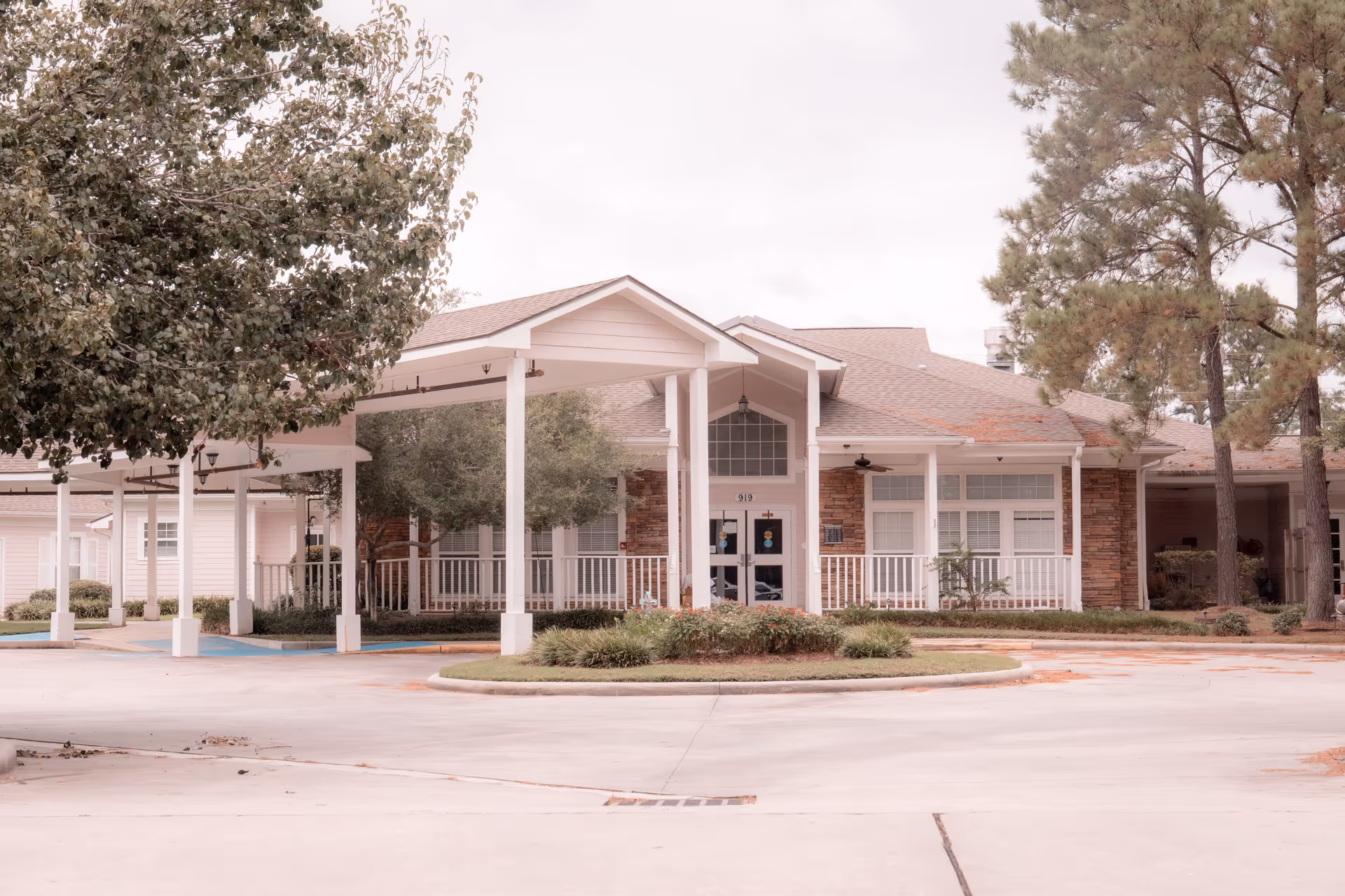 Front exterior view of a single-story building with a covered entrance, white pillars, and a circular driveway. The building has a mix of brick and siding exterior with large windows and is surrounded by trees and landscaping.