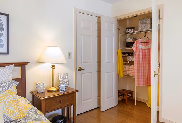 A well-lit bedroom corner with a bed, wooden nightstand and lamp, and an open closet displaying hanging clothes and folded linens.