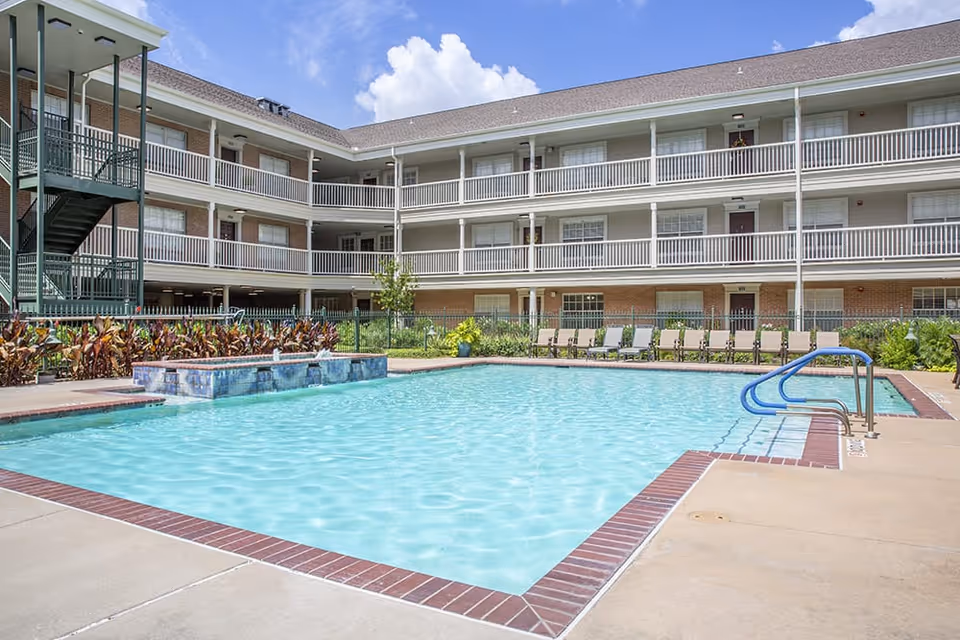 Outdoor swimming pool with clear blue water surrounded by a concrete deck and lounge chairs. In the background, there is a three-story residential building with balconies and a staircase. The sky is partly cloudy and blue.