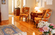 A cozy interior living space with polished hardwood floors, two floral upholstered chairs, a wooden side table with a lamp, a decorative rug, and a bouquet of pink and white flowers in the foreground. The background shows a hallway with a door and a small cabinet with a mirror above it.