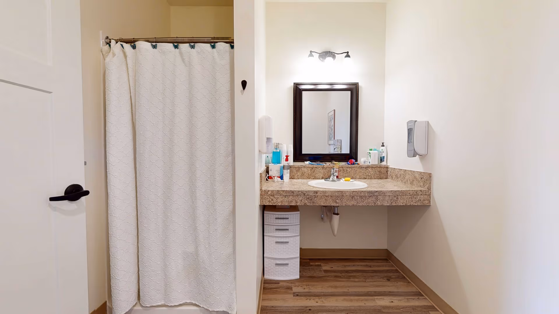 A clean and simple bathroom with a white shower curtain on the left, a countertop with a sink in the center, a mirror above the sink, and various toiletries on the countertop. The walls are light-colored and the floor has wood-like flooring.