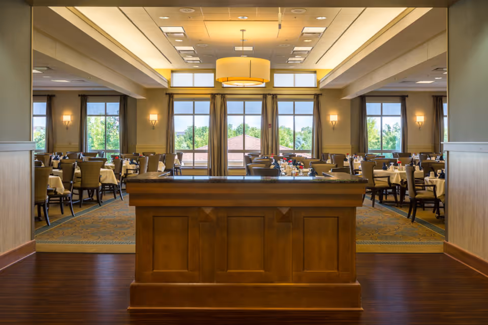 Spacious dining room with a central wooden host stand, neatly set tables and large windows showing greenery outside.