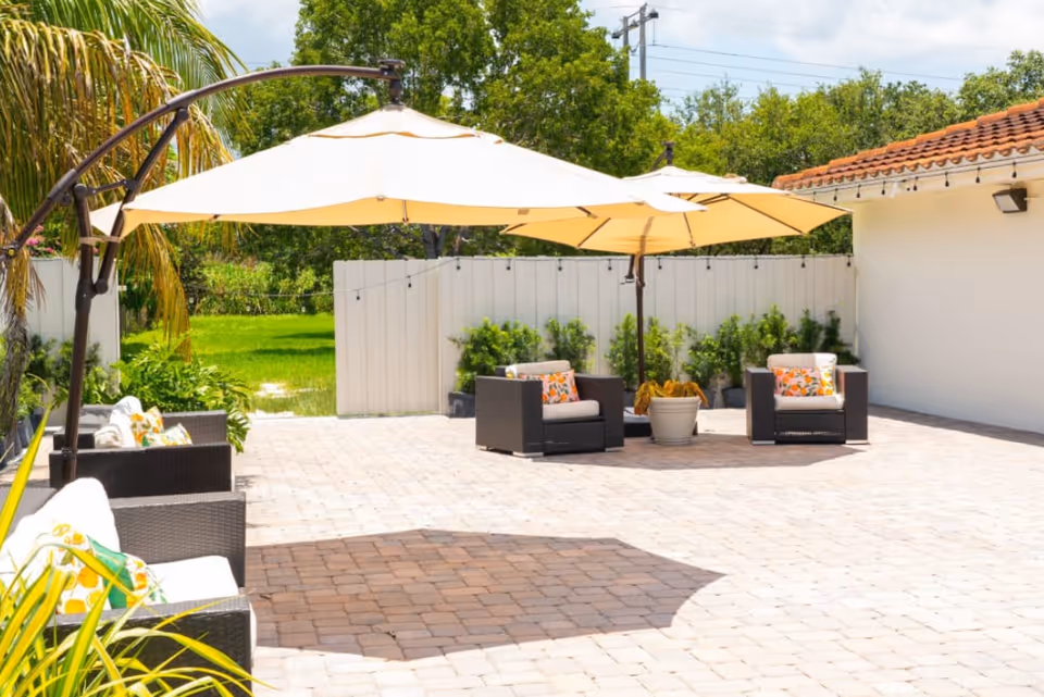 Outdoor patio area with wicker chairs featuring colorful cushions, large beige umbrellas providing shade, potted plants along a white fence, and a building with a tiled roof in the background under a partly cloudy sky.