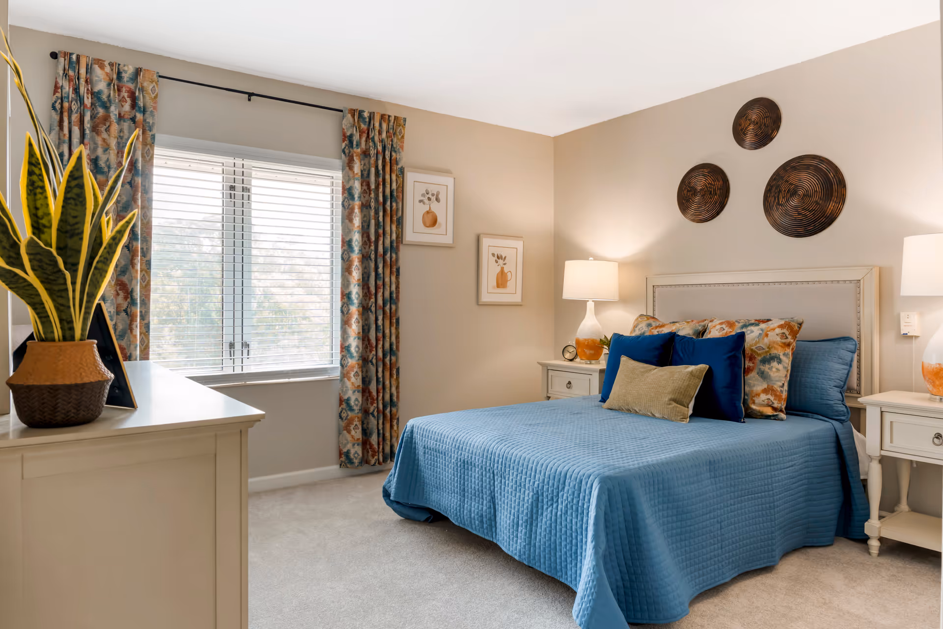 A neatly made bed with a blue quilt and multiple decorative pillows in a bedroom. The room features two bedside tables with lamps, a window with patterned curtains, two framed pictures on the wall, and three circular wall decorations above the bed. A dresser with a potted plant is visible in the foreground.
